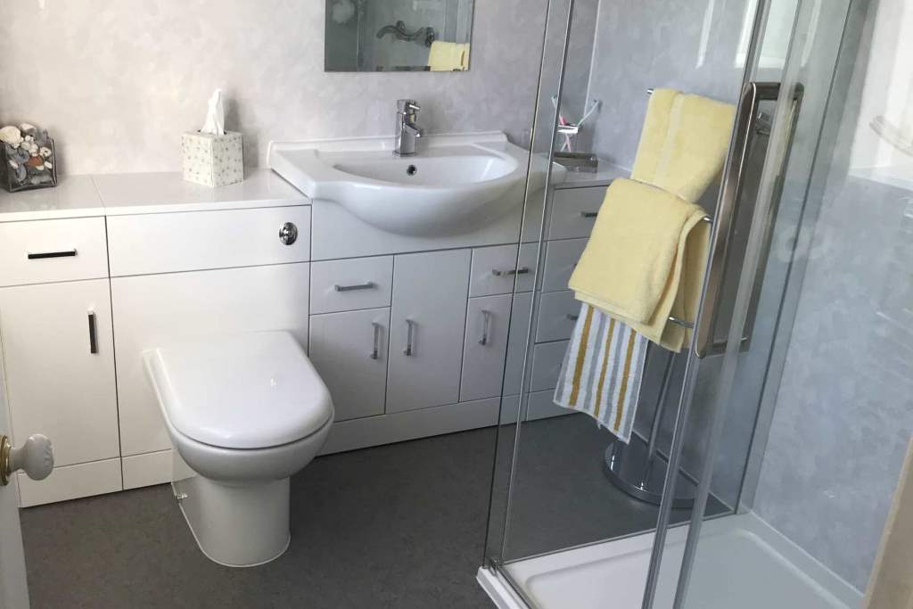 Example of the Type of luxury bathrooms we can install in Birmingham homes - shows modern white bathroom suite with sink and toilet embedded into wall of cabinets and a walk in shower with whole room sealed as wet room